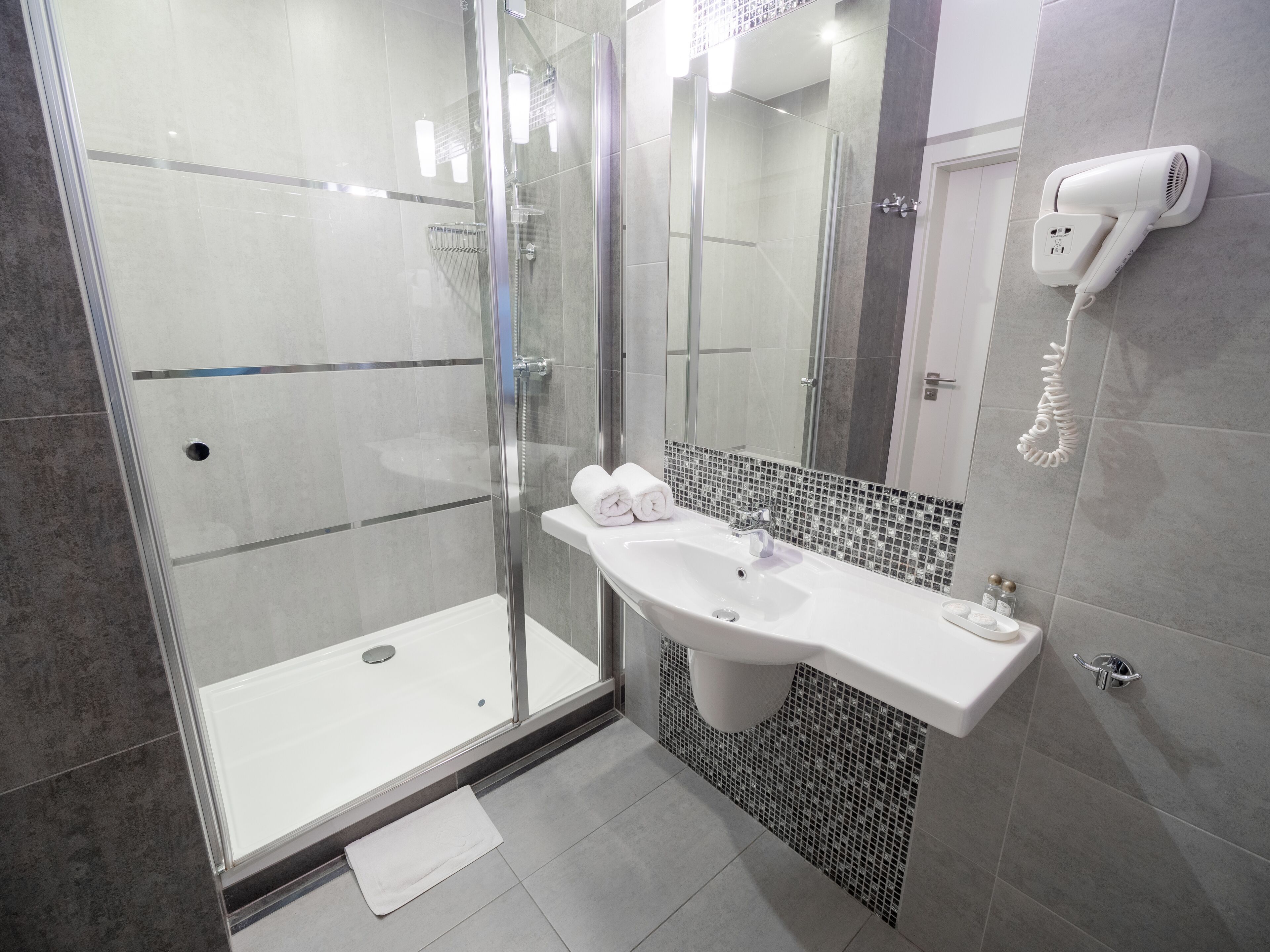 A modern bathroom with a glass-enclosed shower, a white sink with a mirror above it, and a hairdryer mounted on the wall.