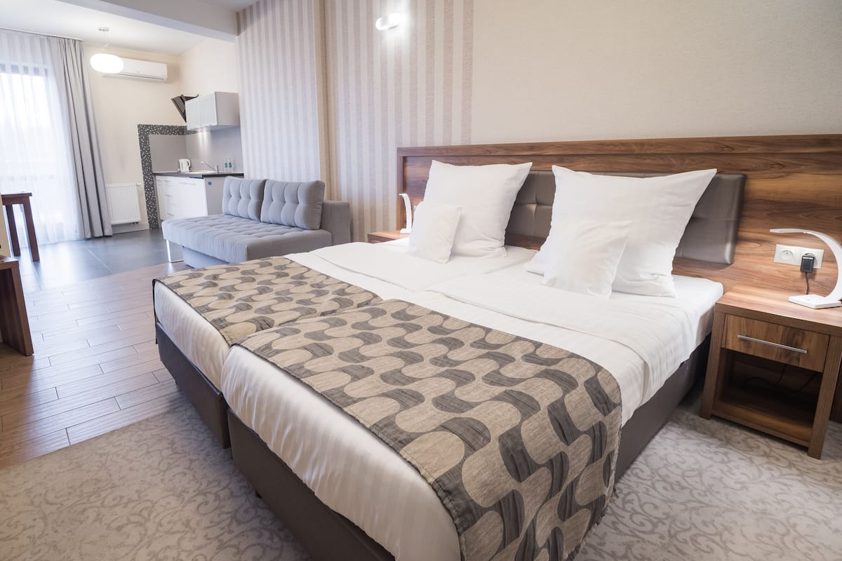 Quarto de hotel moderno com uma cama grande, uma mesa de cabeceira de madeira e uma pequena mesa ao lado da cama, além de vista para a área de estar adjacente.