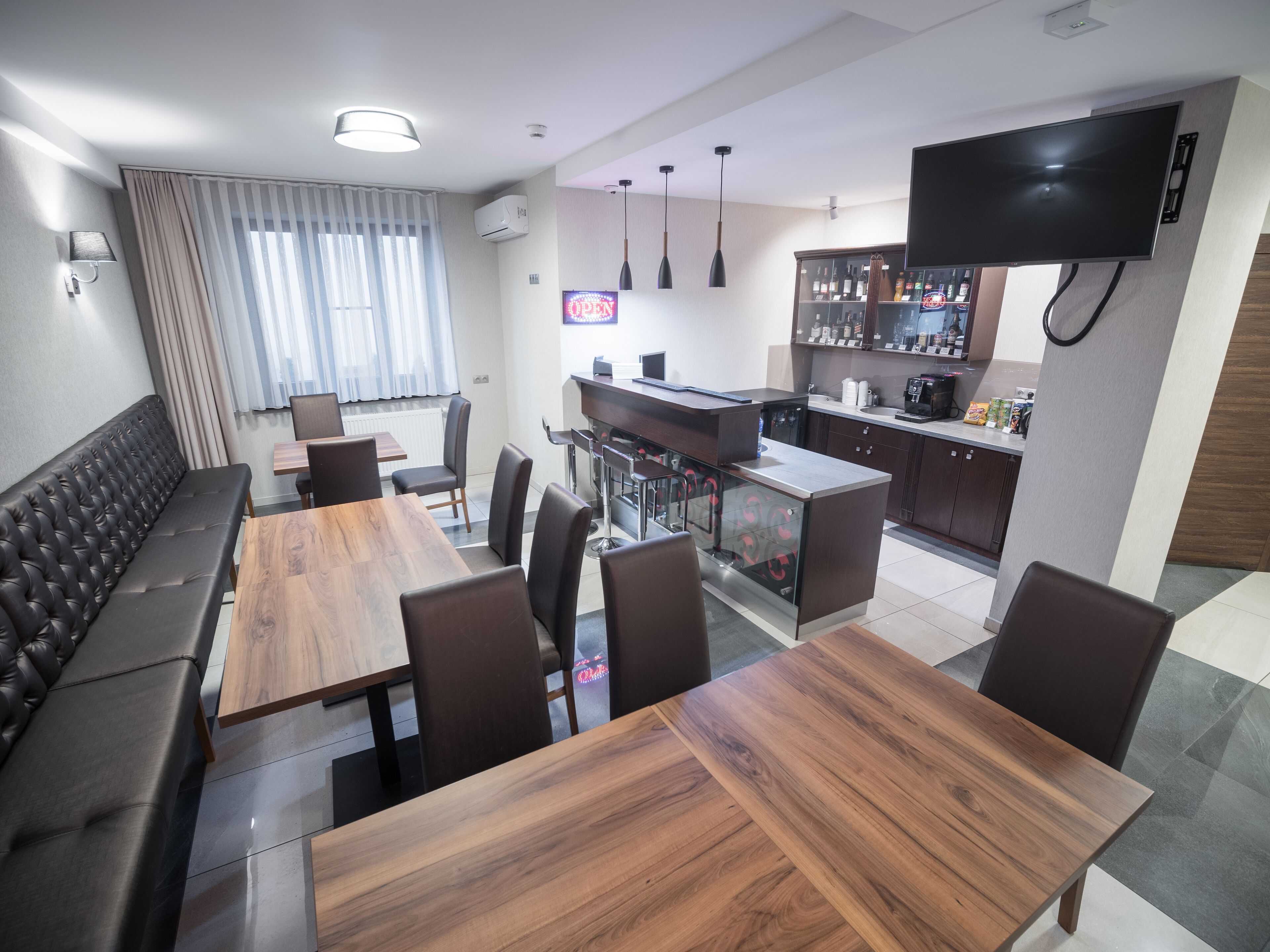 A modern dining area with a long wooden table and cushioned seating, a kitchen area with a bar, and a flat-screen TV mounted on the wall.