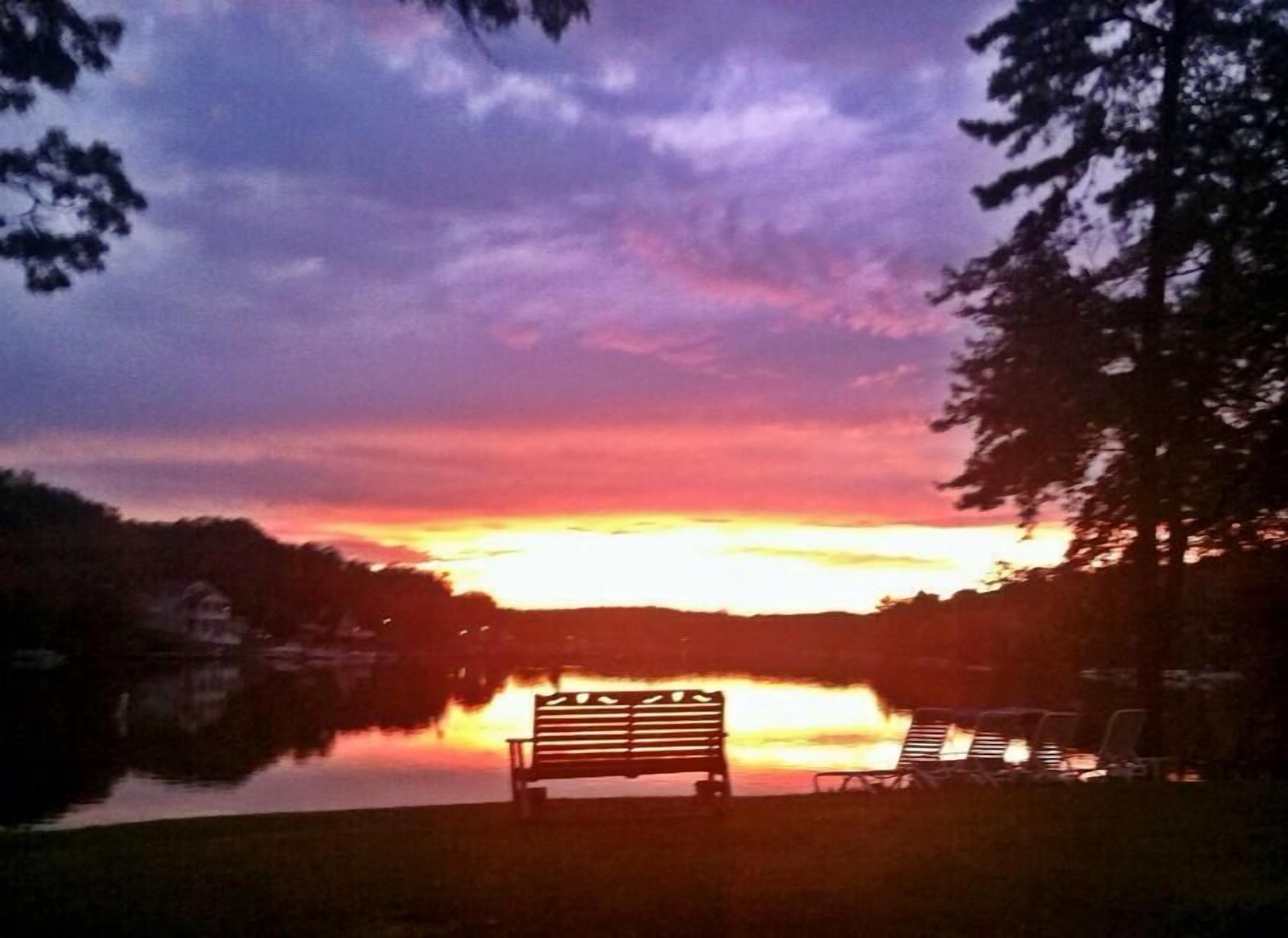 A bench and lounge chairs by a lake at sunset.