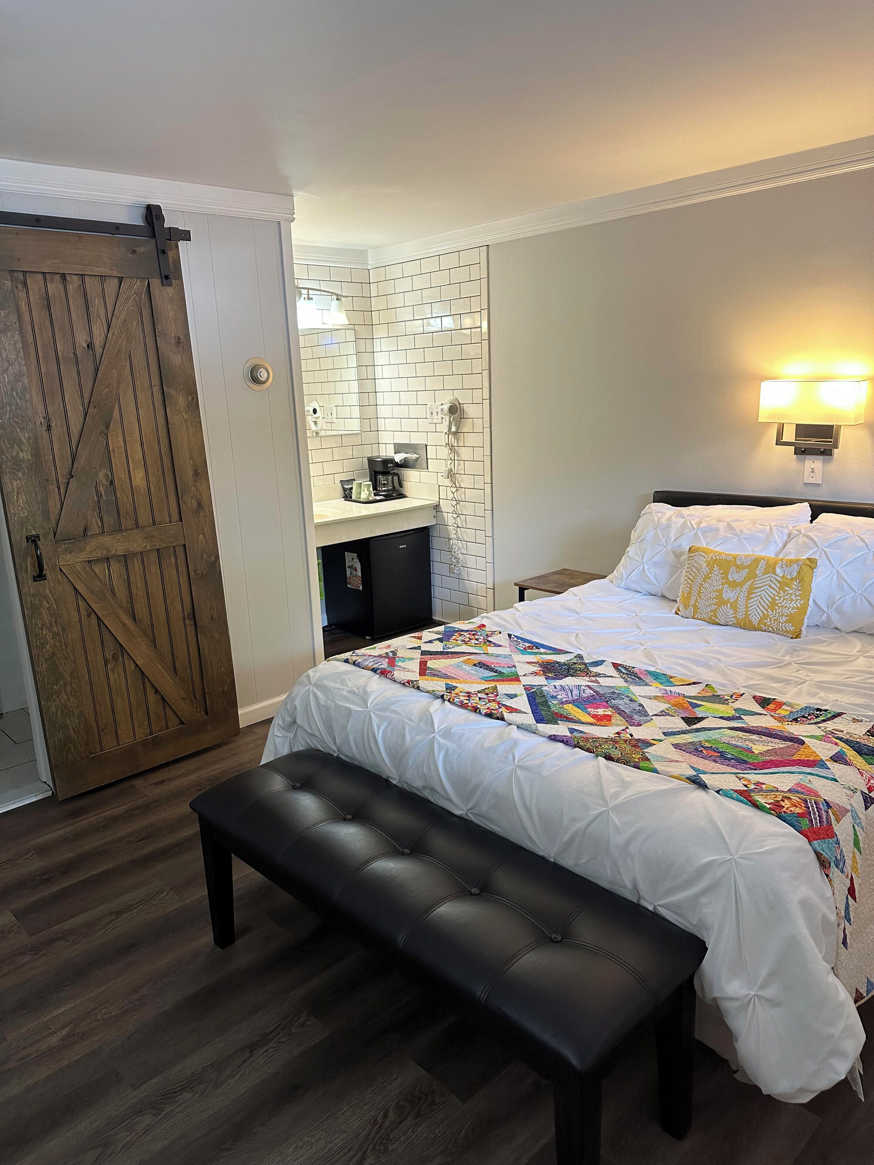 A bedroom with a large bed, a bench, a wooden door, a kitchenette, and a wall-mounted light fixture.