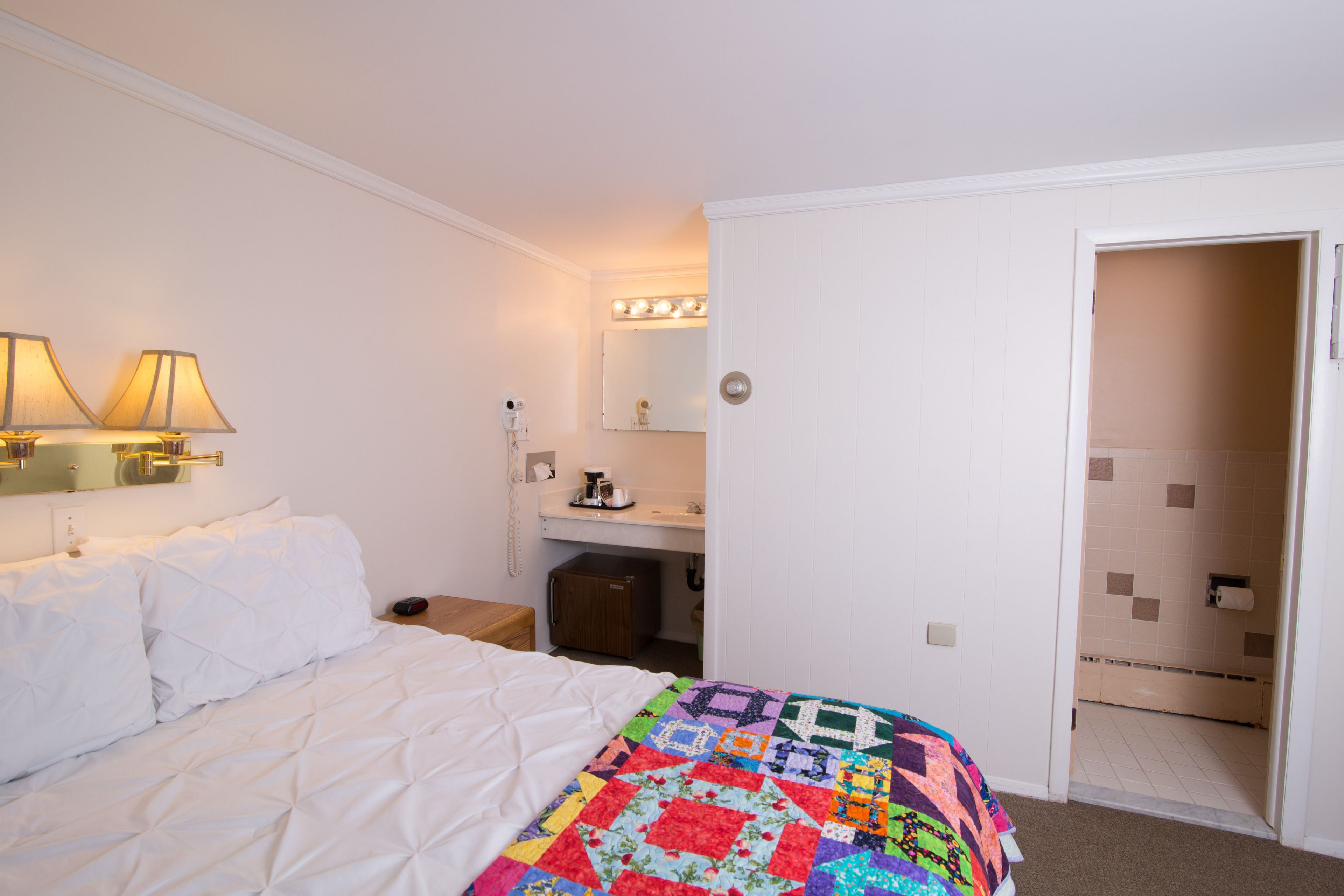 A hotel room with a neatly made bed, a colorful quilt, a bedside lamp, a small table with a coffee maker, a mirror with lights, and a door leading to a bathroom.