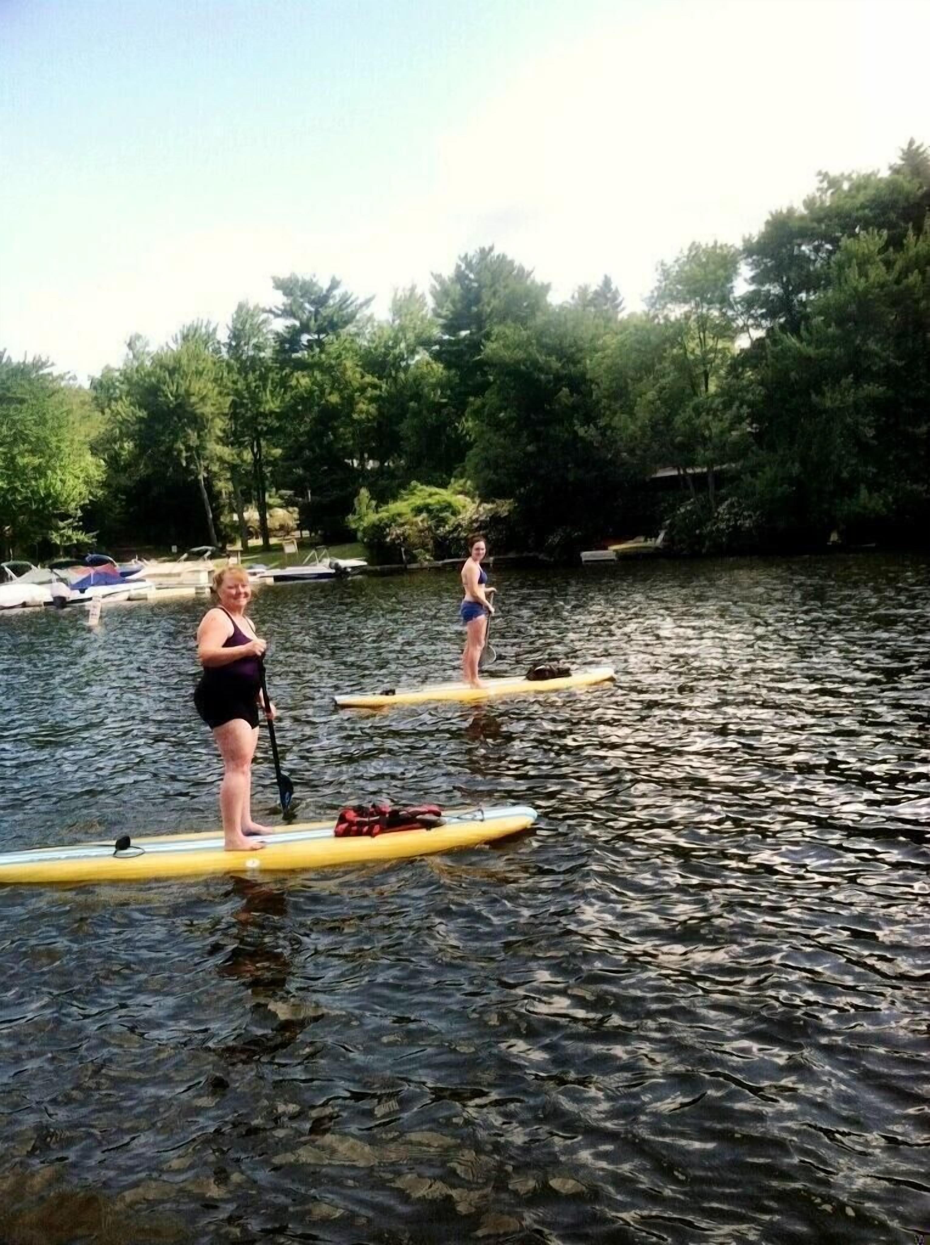 Two people on paddleboards in a calm river with boats and trees in the background.