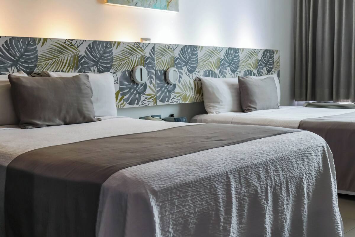 Une chambre d’hôtel avec deux lits, une tête de lit ornée d’un motif de feuilles, un tableau représentant une méduse et une fenêtre avec des rideaux.