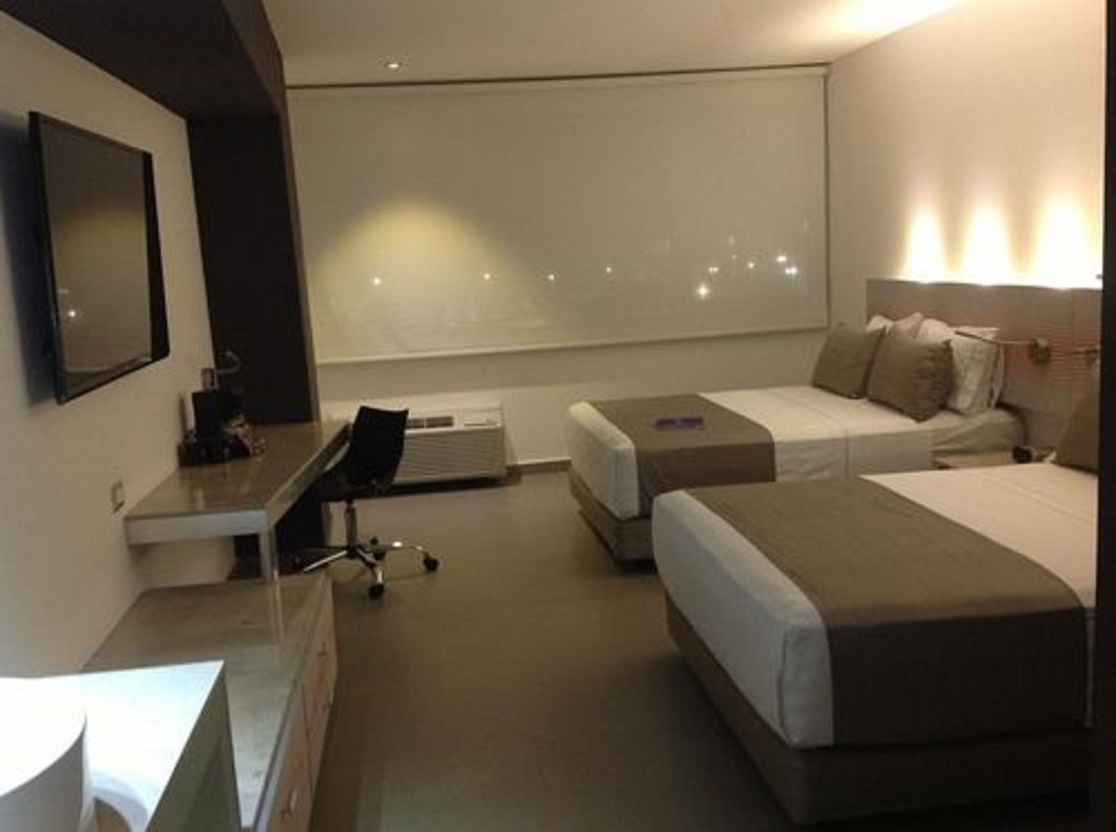 A hotel room with a desk, two beds, a chair, and a large window.