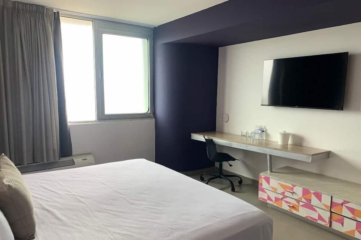 Une chambre d’hôtel avec un lit, un bureau, une chaise, une télévision et une fenêtre avec des rideaux.