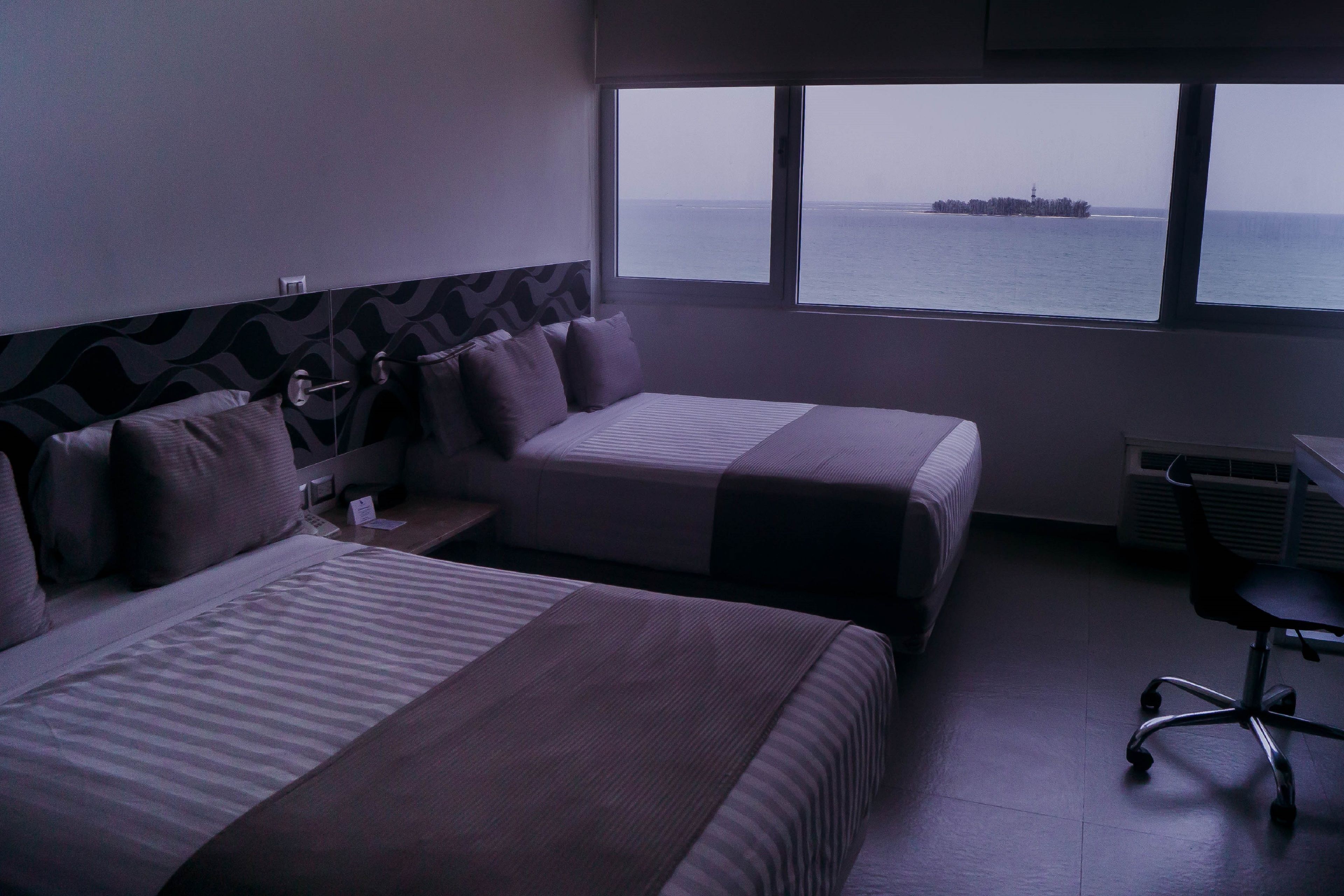 A hotel room with two beds, a desk, and a view of the sea.
