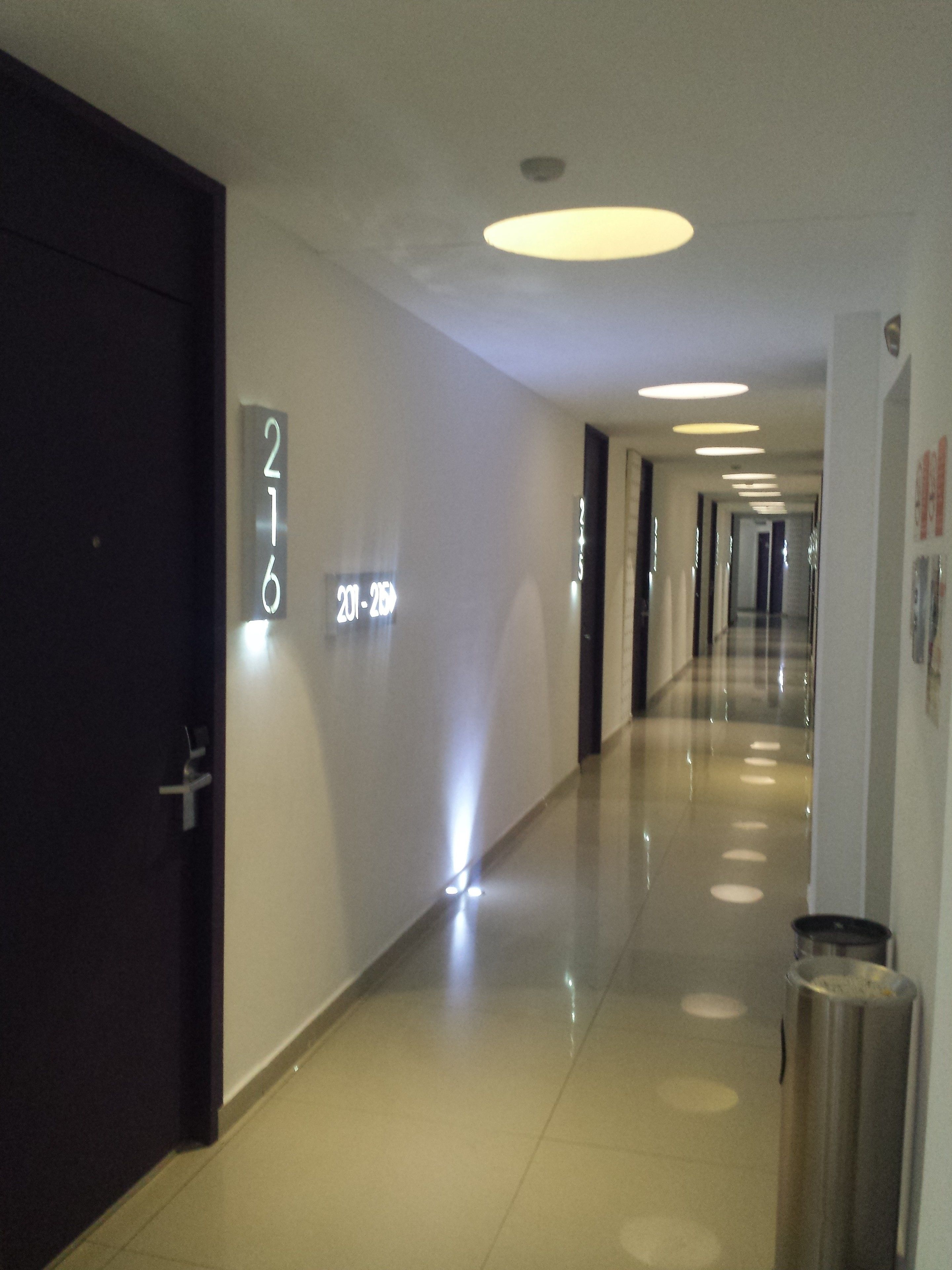 A modern hotel corridor with a clean, reflective floor, a trash bin, and illuminated wall numbers indicating room 216.