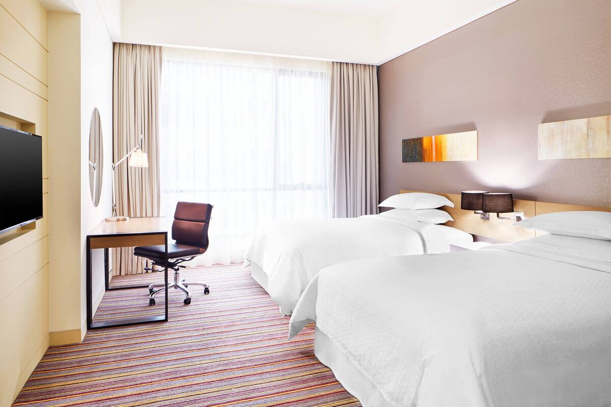 Ein Hotelzimmer mit zwei Betten, einem Schreibtisch mit Stuhl, einem Fernseher und einem großen Fenster mit Vorhängen.