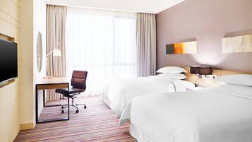 Habitación de hotel con dos camas, un escritorio con silla, televisión y un ventanal con cortinas.