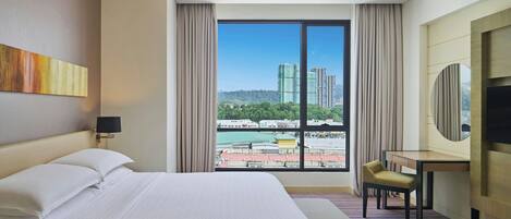 Habitación de hotel con una cama grande, un escritorio, una silla, un televisor y vistas a edificios y zonas verdes.