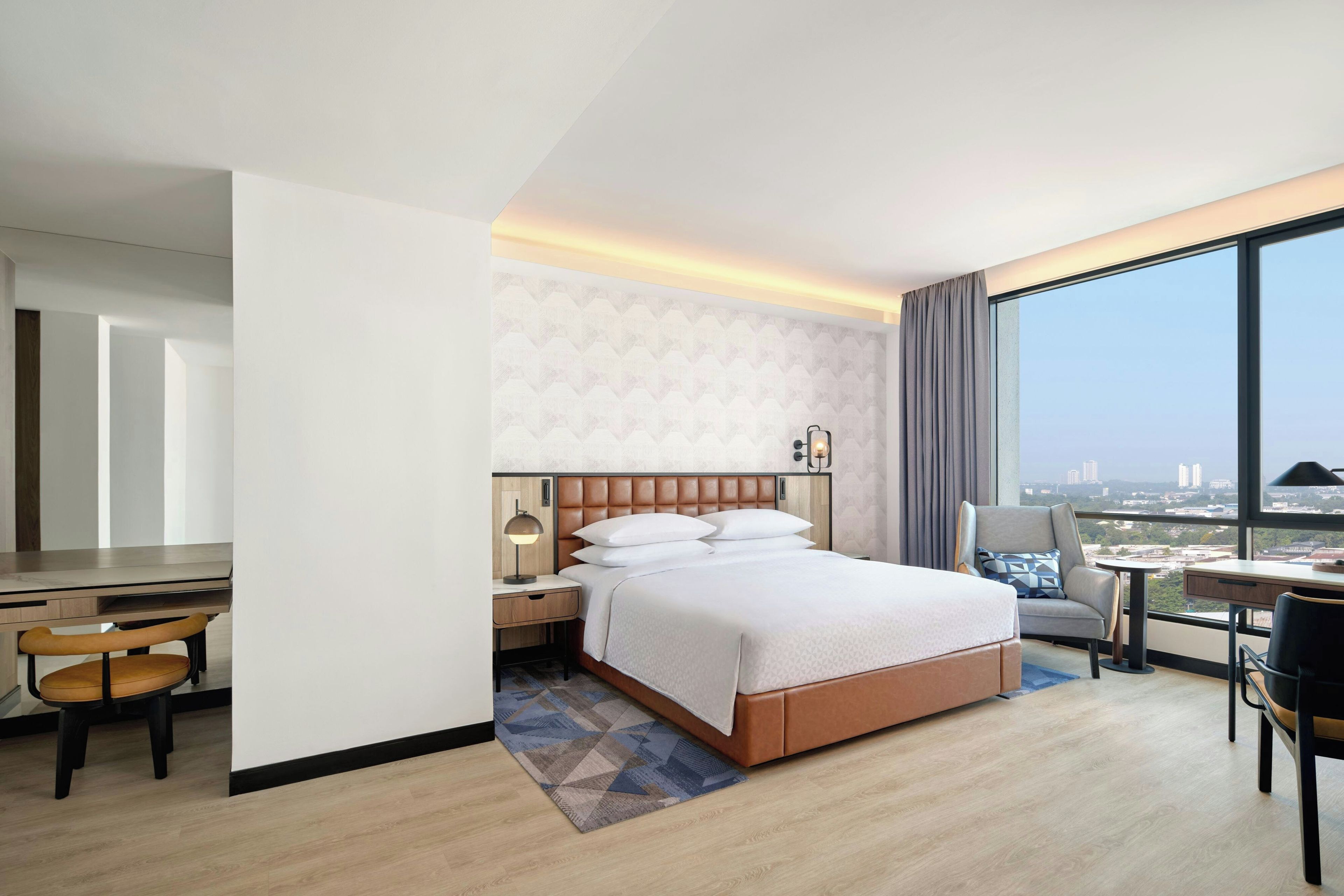 A hotel room with a bed, a desk, a chair, a nightstand, and a view of the city.