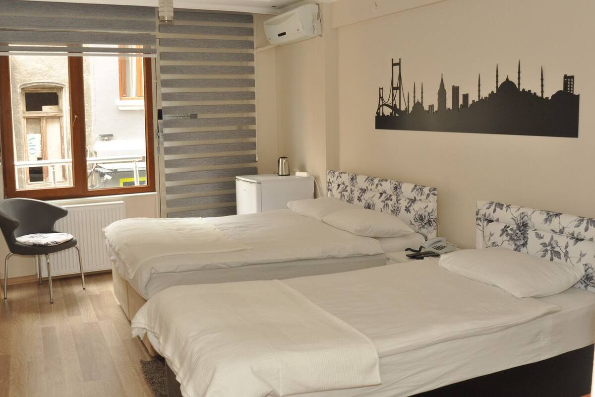 A hotel room with two beds, a chair, a radiator, a kettle, and a cityscape wall art.