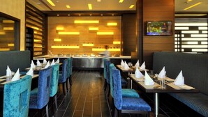 A restaurant with a modern interior, featuring blue upholstered chairs, a central bar area, and a television mounted on the wall. - Hotel Marigold Jaipur (Jaipur)