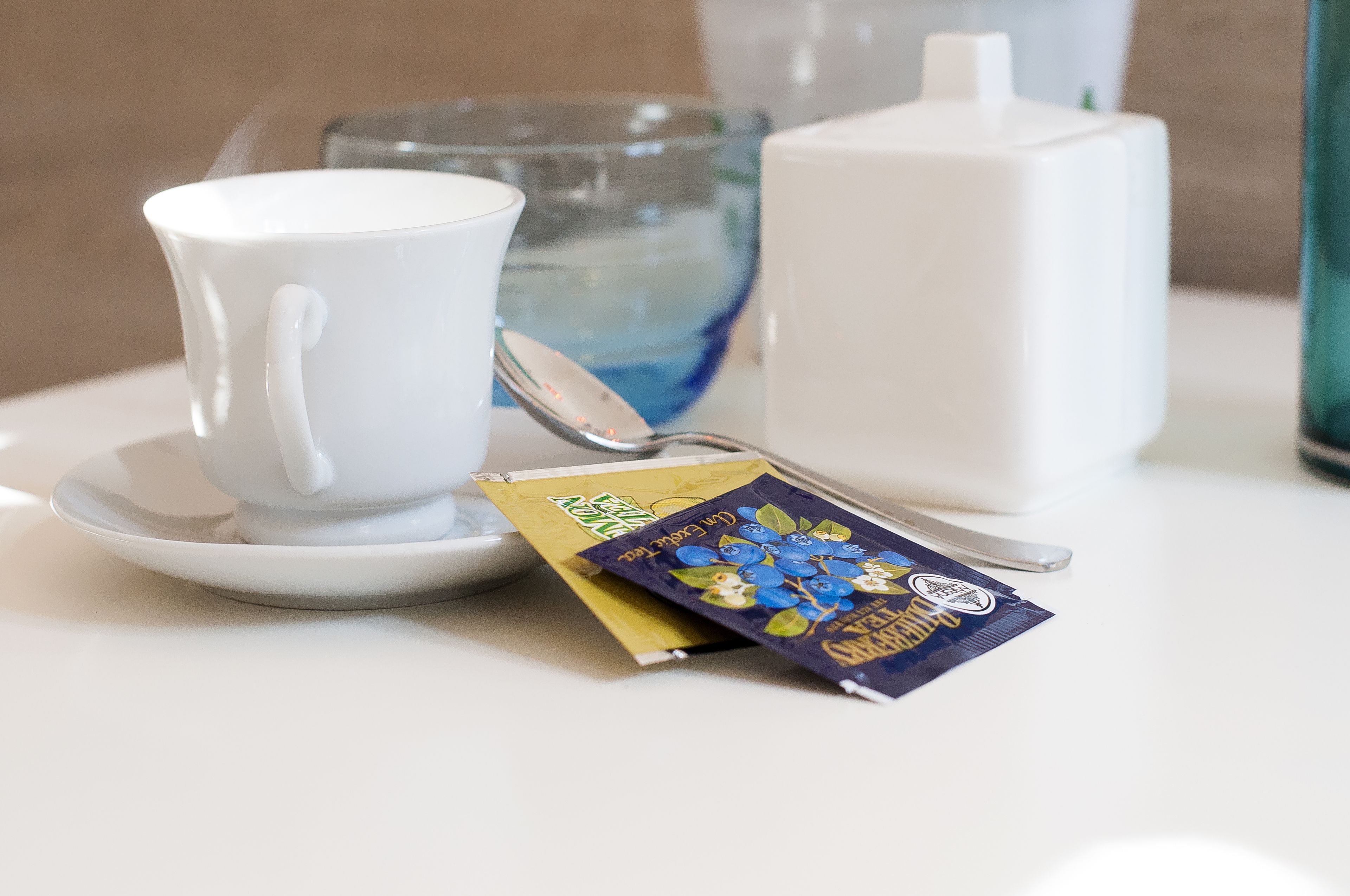 Une tasse blanche posée sur une soucoupe, un verre d’eau, un récipient blanc et une boîte de thé dont l’étiquette est bleue et jaune.
