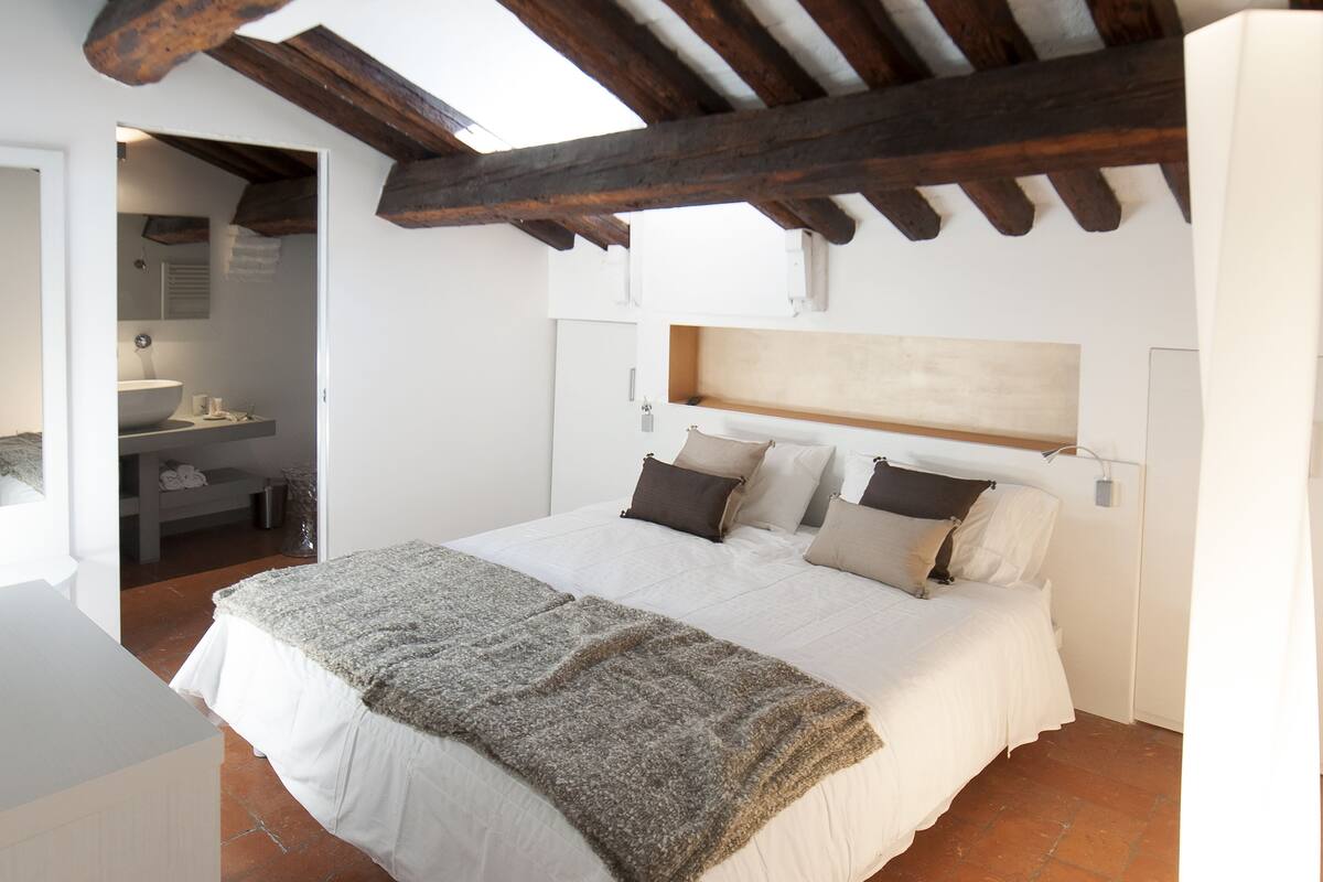 A bedroom with a large bed, wooden beams, a white headboard, and a view of a bathroom.