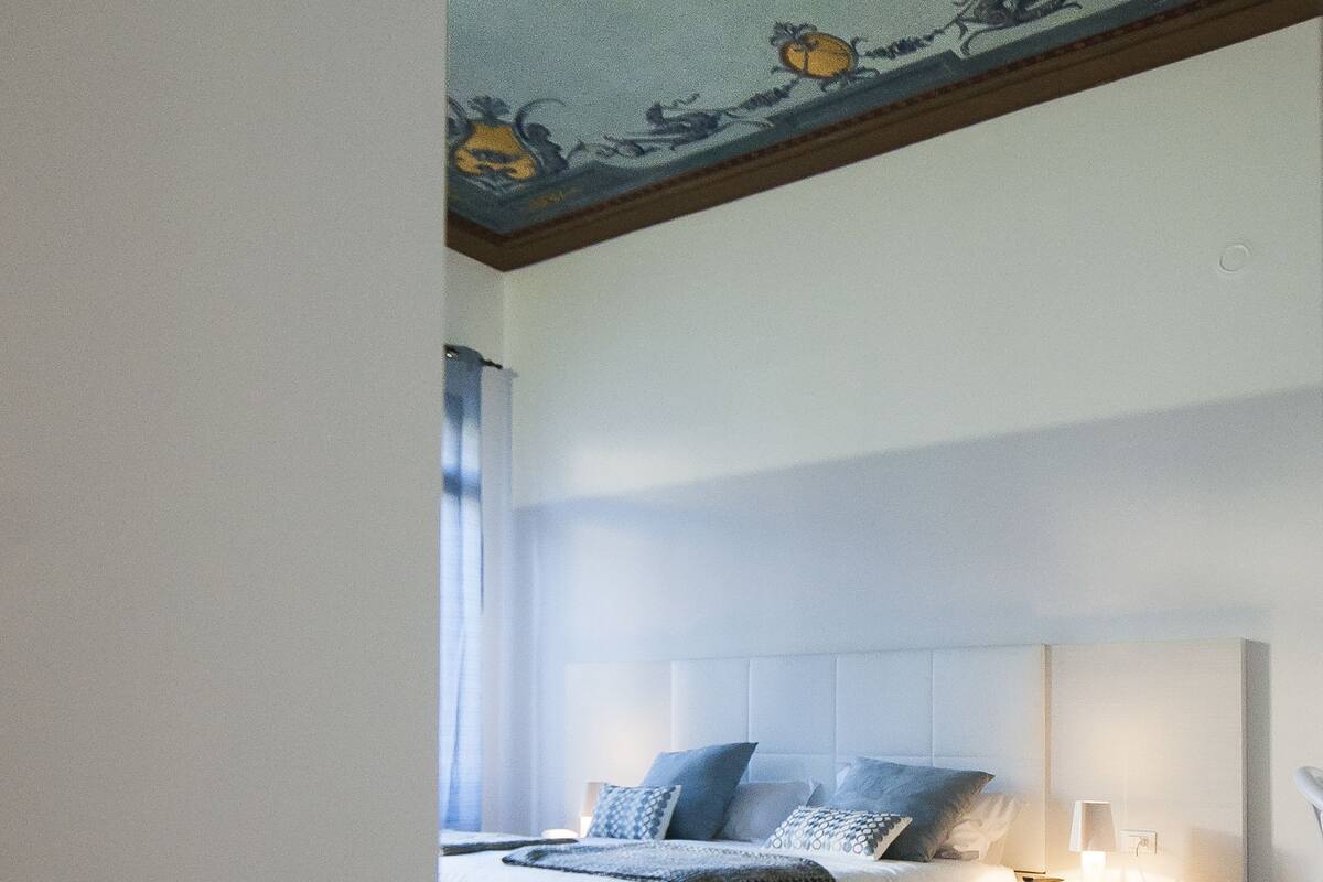 A bedroom with a wooden floor, a bed with white bedding, a nightstand, and a ceiling with floral and fruit designs.
