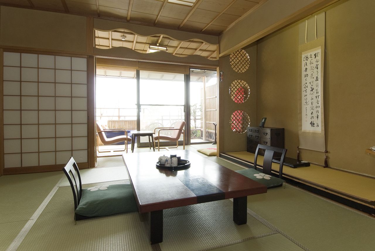 Ein traditioneller japanischer Raum mit Tatami-Böden, einem niedrigen Tisch mit Kissen und Schiebetüren, die auf einen Balkon führen.