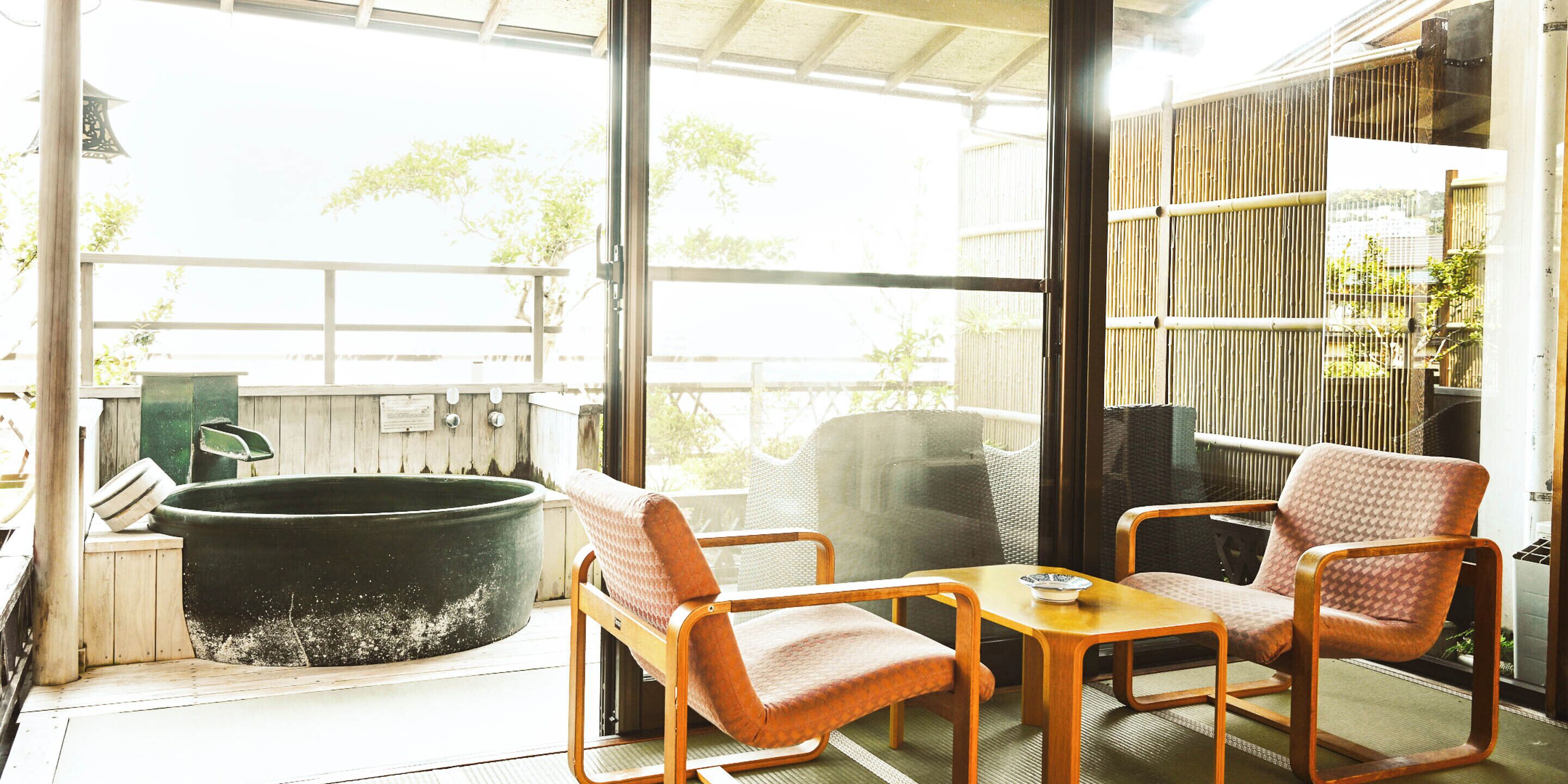 A room with a large black bathtub, two wooden chairs, a small table, and a view of greenery outside.