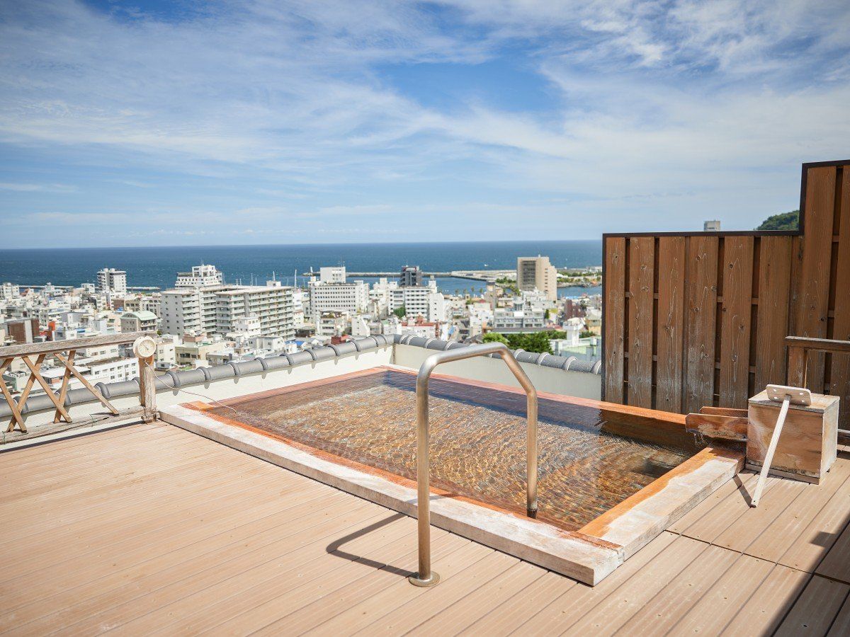 A rooftop pool with a wooden deck, metal railings, and a view of the city and ocean.