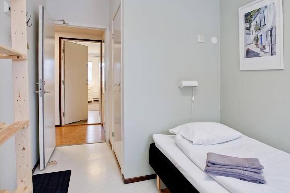 A small, single-bed room with a wooden floor, a white wall, and a door leading to another room.