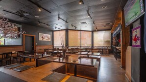 A restaurant with wooden tables and benches, large windows with blinds, and a sports screen on the wall. - Cedars Inn Hotel & Convention Center (Gibsons)