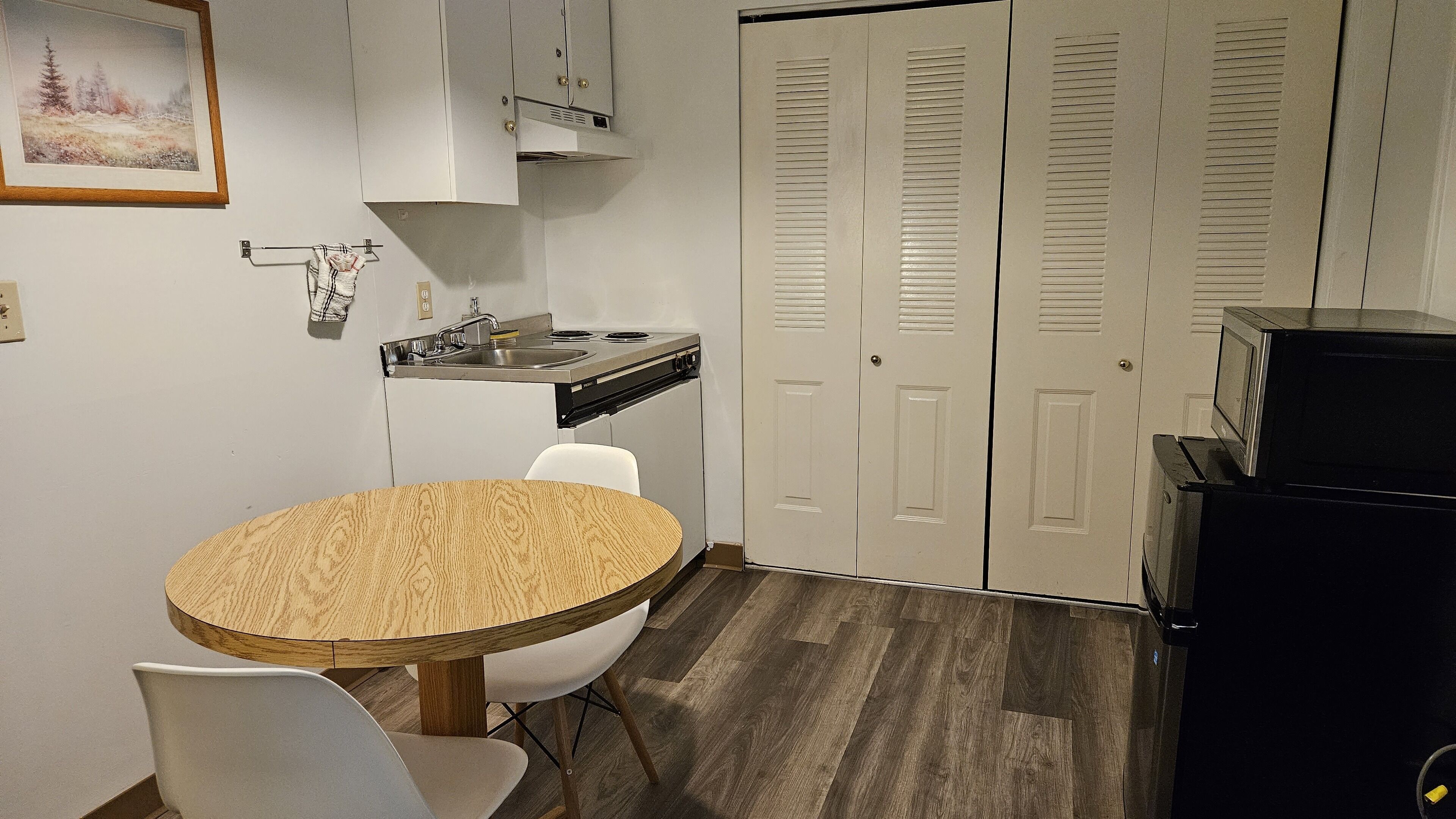 A compact kitchen with a round wooden table, white chairs, a stove, and a microwave. There are two white doors with louvered windows.