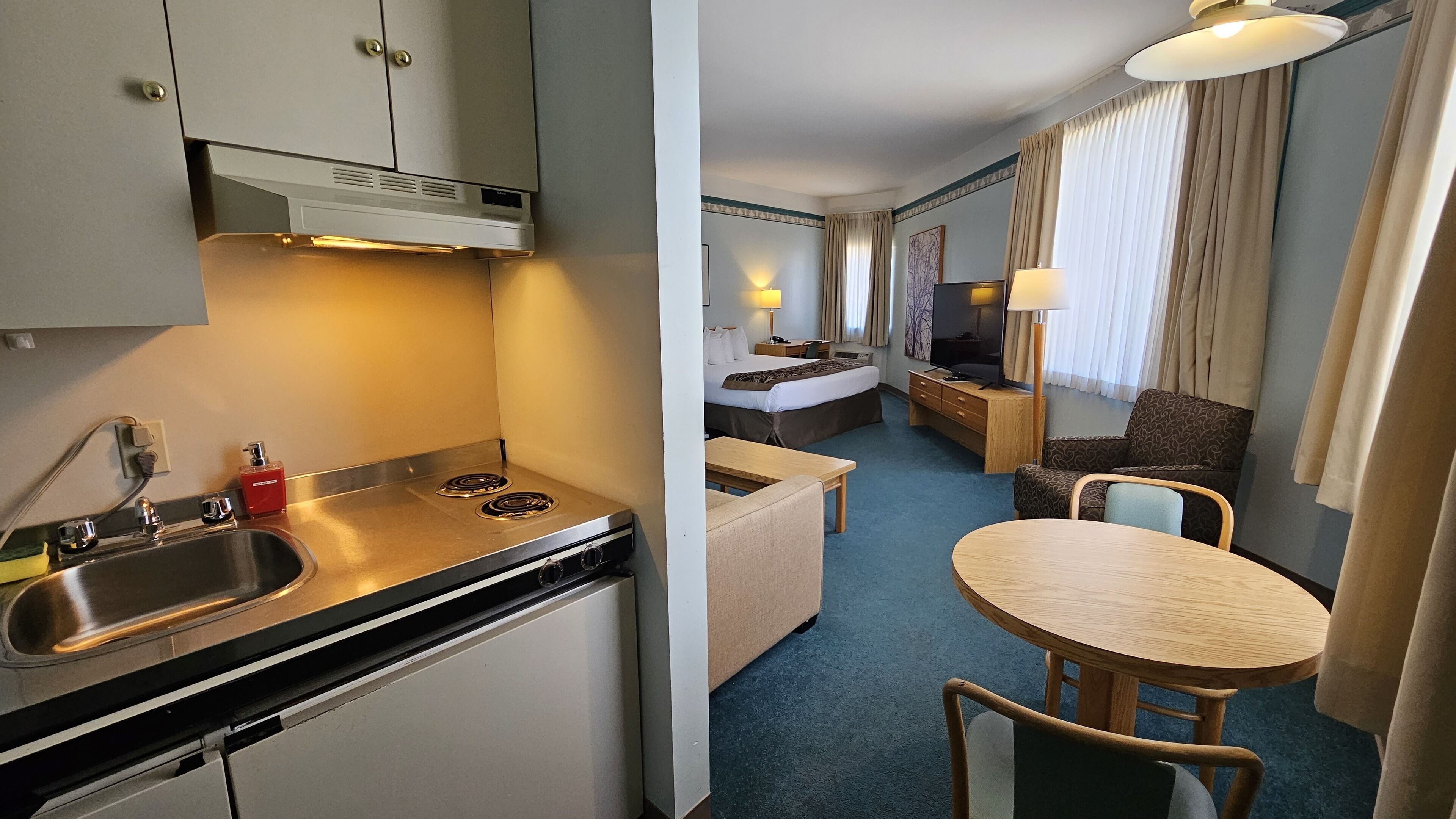 A compact hotel room with a kitchenette, a bed, a small desk, a TV, and a seating area.
