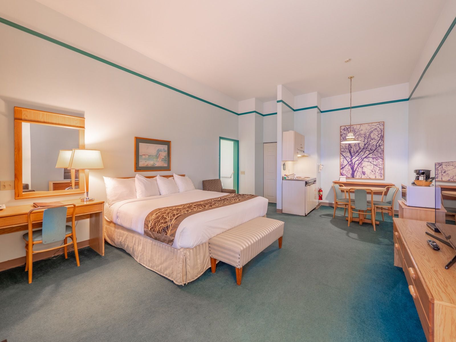 A hotel room with a large bed, a desk, a chair, a small kitchenette, and a dining area with a table and chairs.