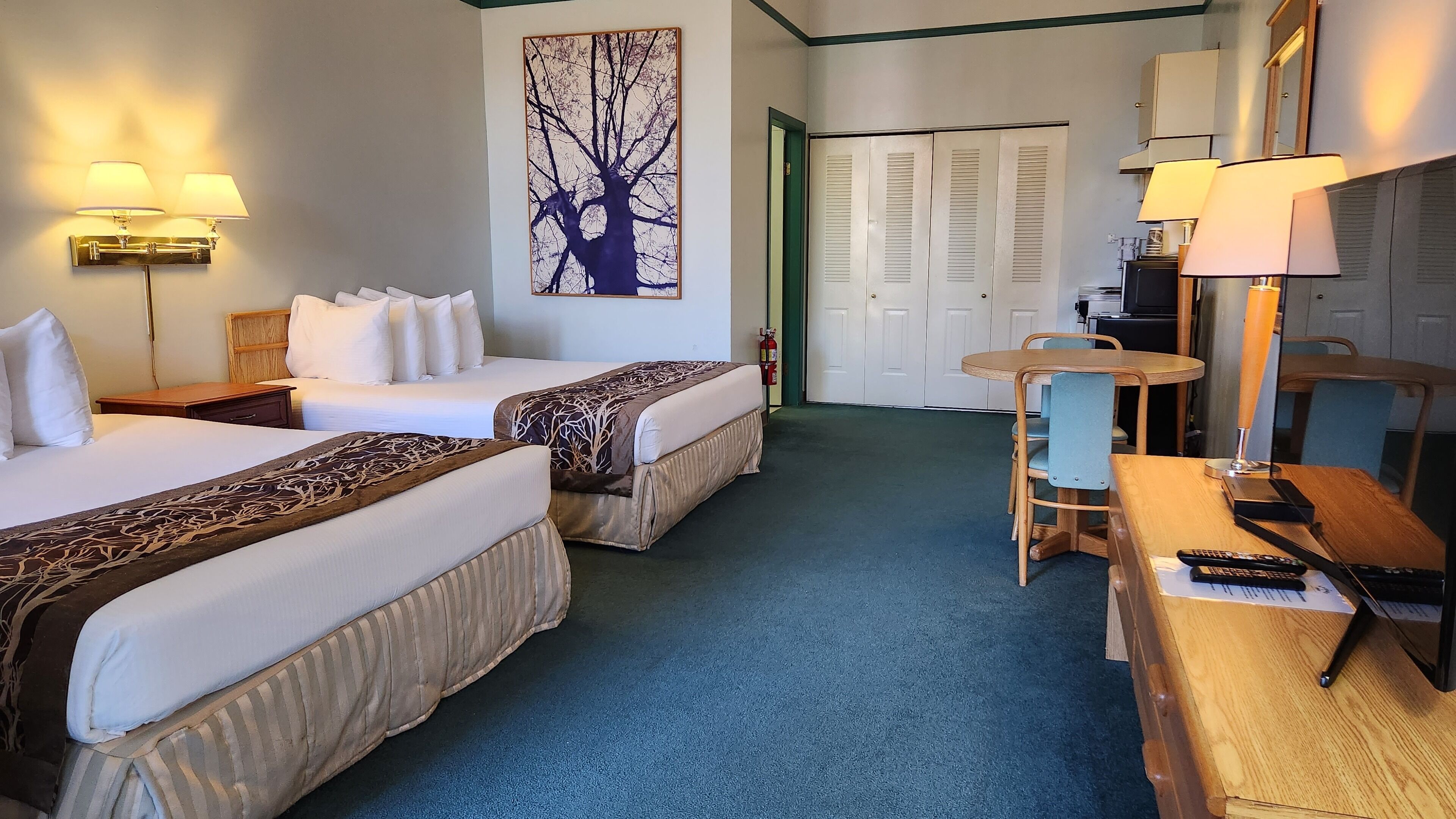Hotelkamer met twee bedden, een eettafel met stoelen, een kleine televisie en een schilderij van een boom aan de muur.