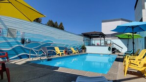 A swimming pool with lounge chairs and umbrellas, surrounded by a building with a blue exterior and a lifeguard sign. - Cedars Inn Hotel & Convention Center (Gibsons)