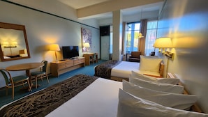A hotel room with two beds, a desk with a TV, a chair, a small table, and a window with curtains. - Cedars Inn Hotel & Convention Center (Gibsons)