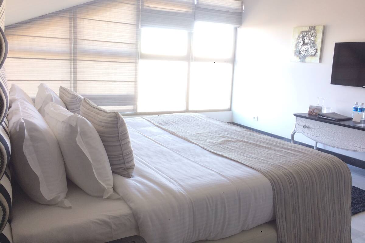 Ein Schlafzimmer mit einem großen Bett, einem Schreibtisch mit Flachbildfernseher und einem Fenster mit Jalousien.