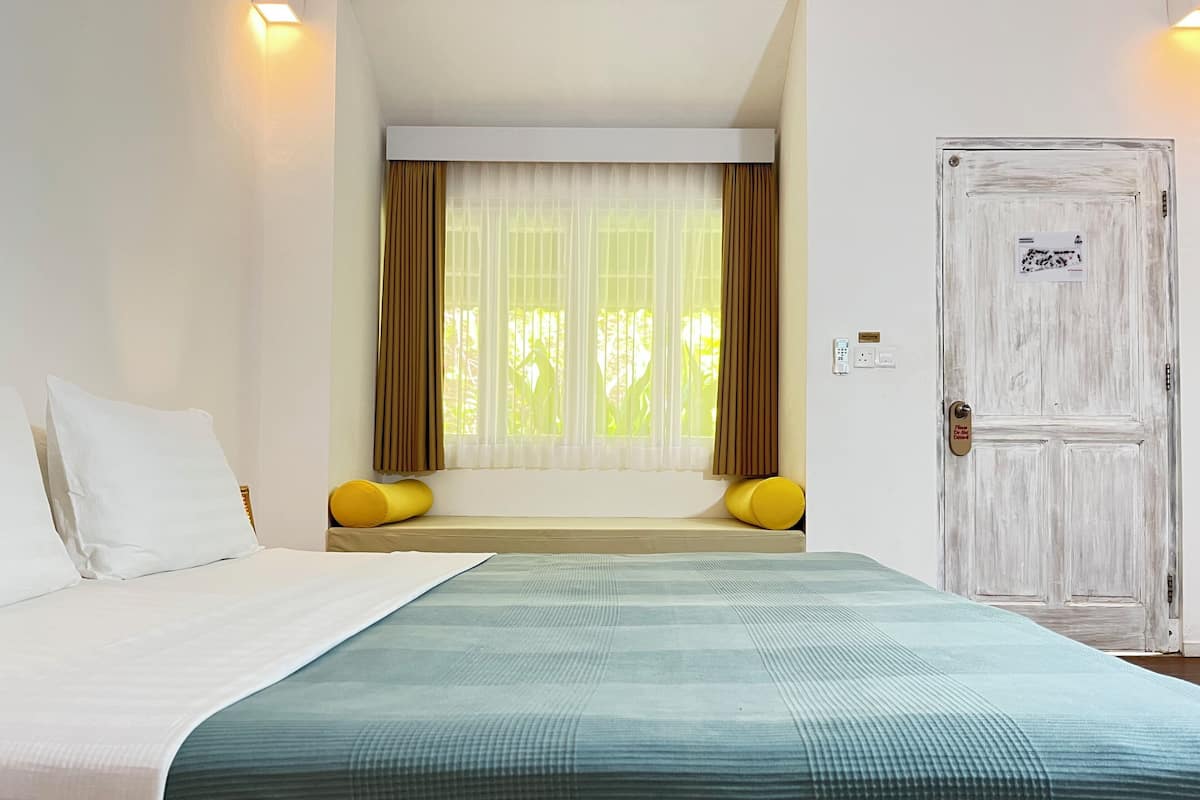 A hotel room with a bed, a window with curtains, a door, and a wall-mounted light.