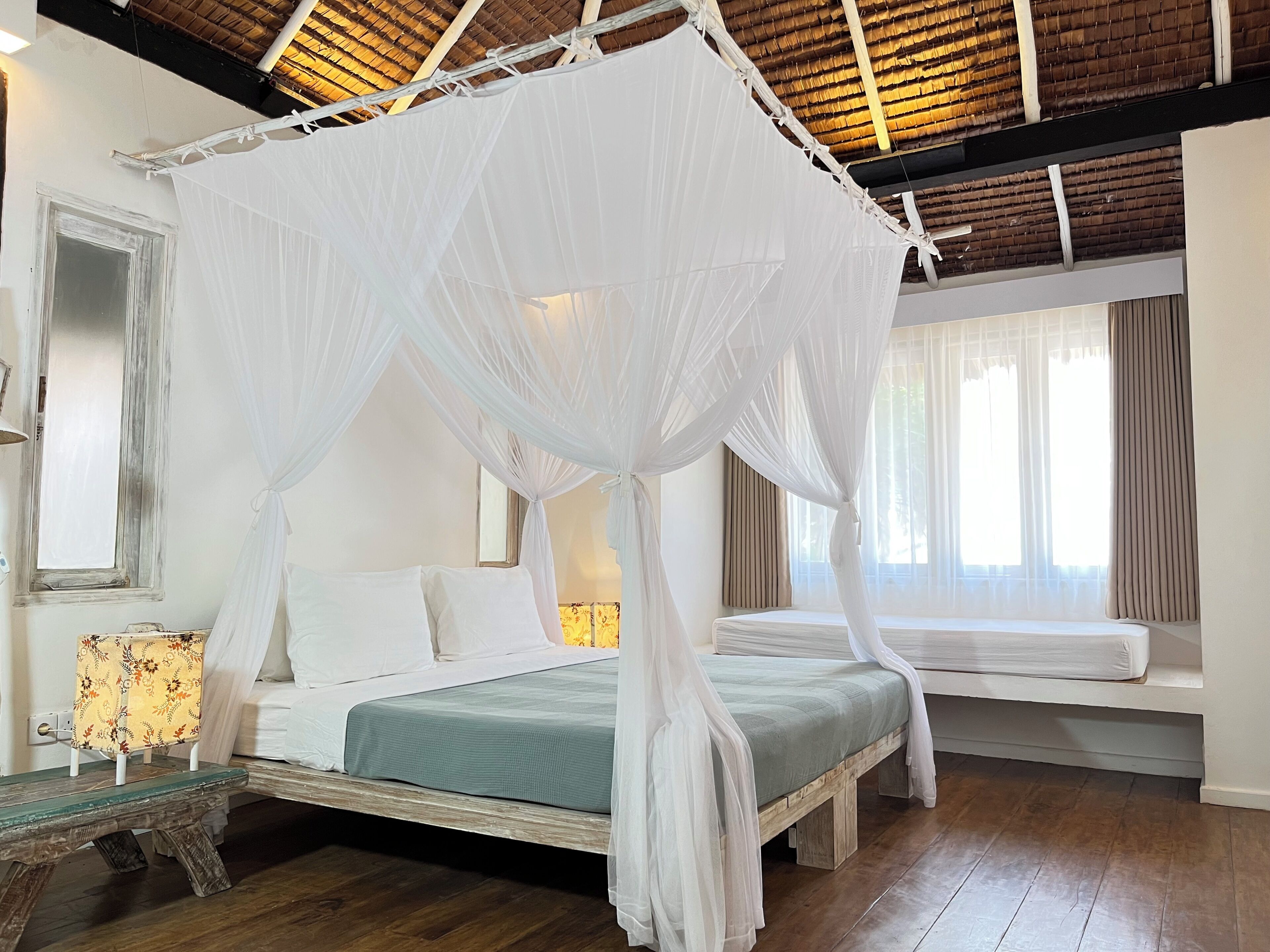 Ett rum med en säng under ett himmelstält, en bänk, ett sängbord och ett fönster med gardiner.