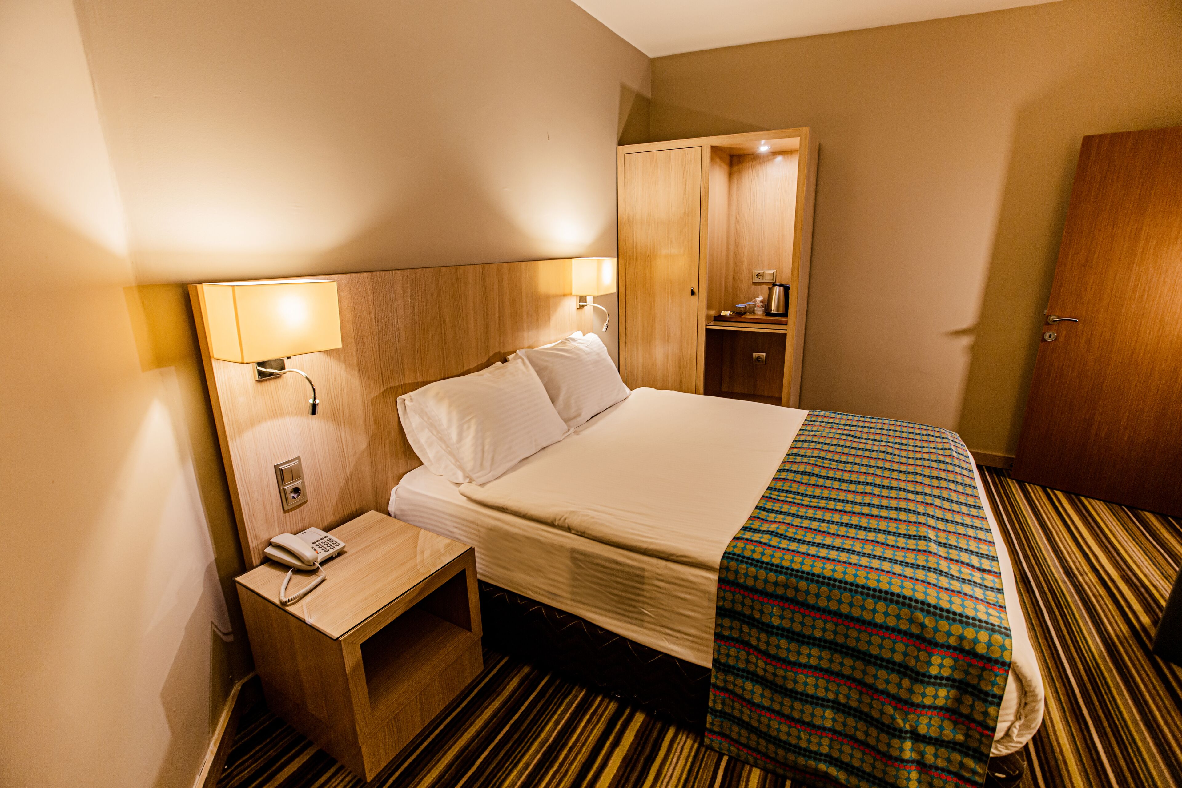 Una habitación de hotel con cama, mesita de noche, lámpara, armario y teléfono.