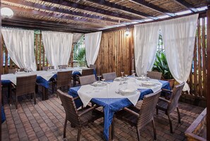 An outdoor dining area with wicker tables and chairs, covered by a wooden pergola and white curtains. - Hotel Mannu (Bosa)