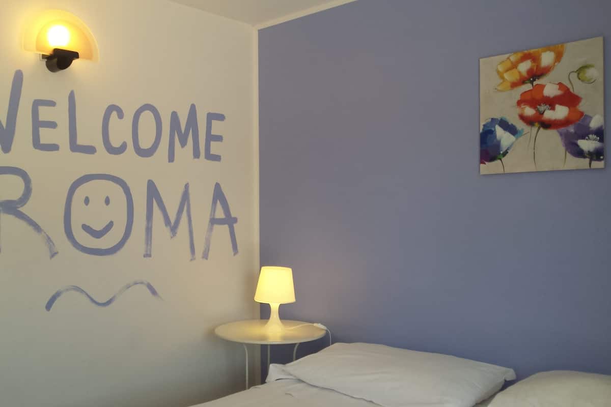 A room with a wall sign reading "WELCOME ROMA" and a painting of flowers.