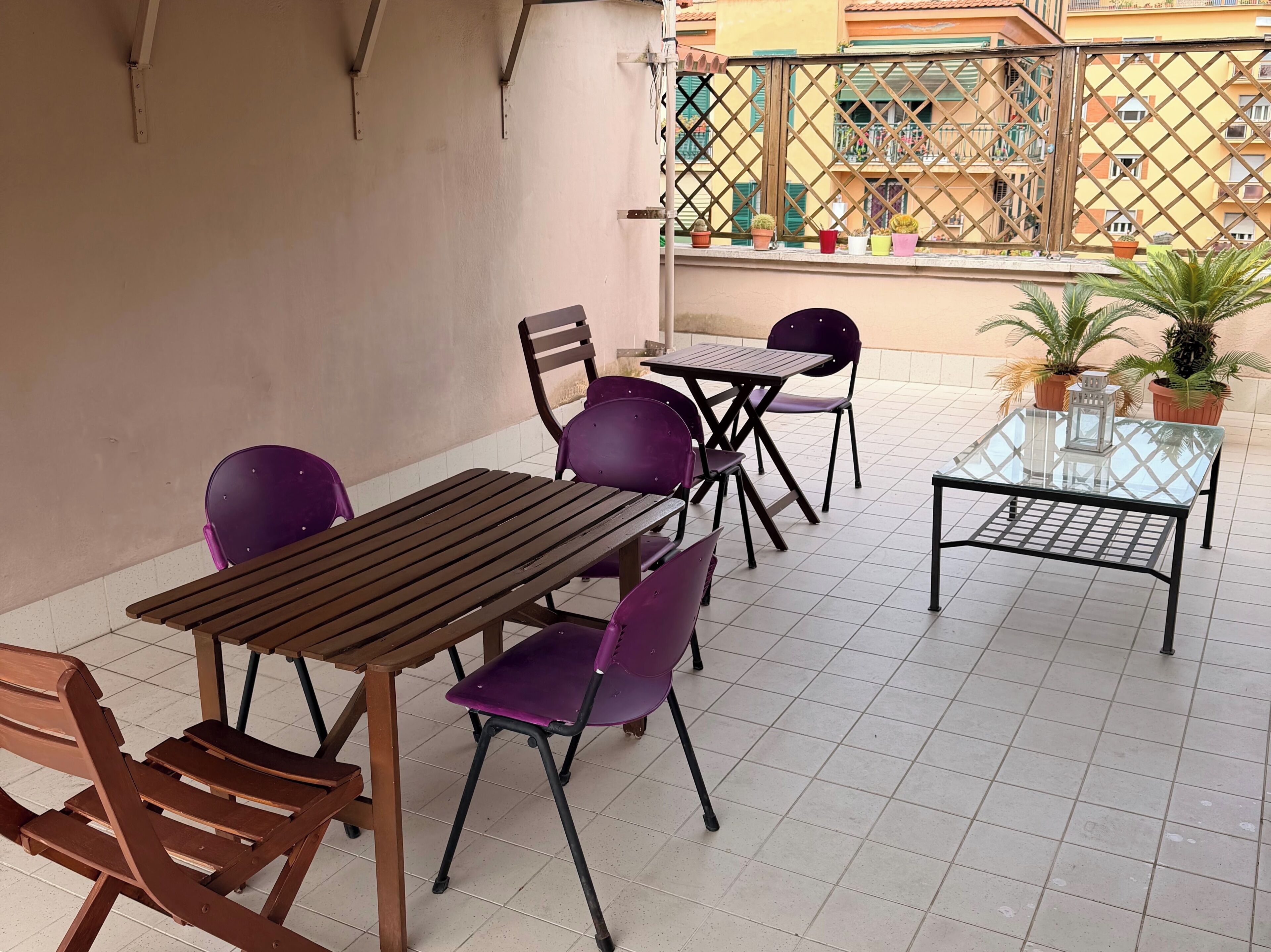 A terrace with a wooden table and purple chairs, a glass table with a metal frame, and potted plants.