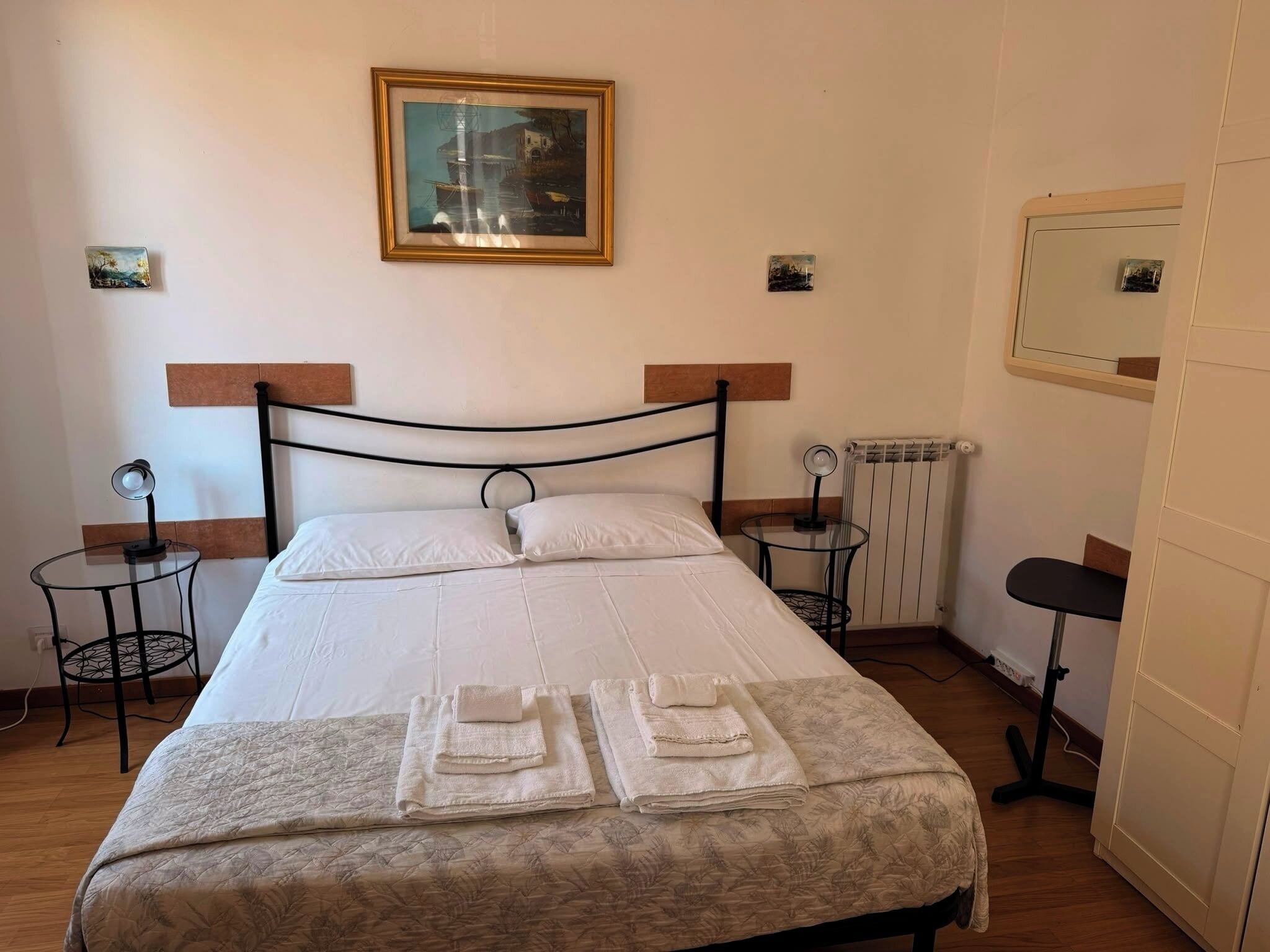 A neatly made bed with a white duvet and two folded towels, a small round table with a lamp, and a radiator on the wall.