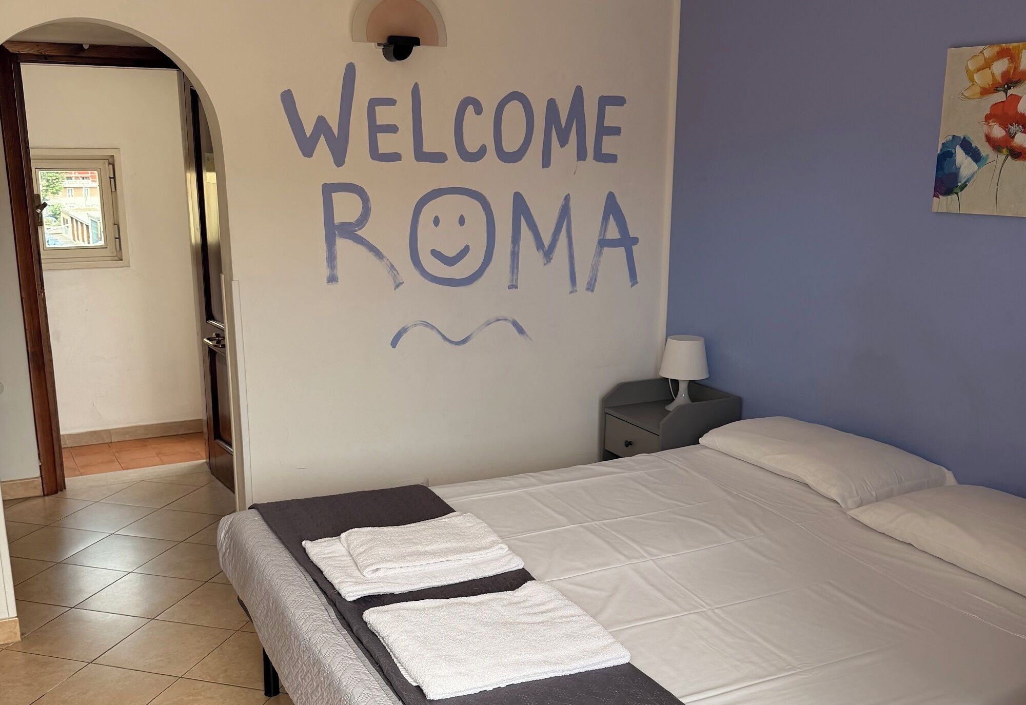 A room with a bed, a nightstand, a lamp, and a wall with 'WELCOME ROMA' written on it.