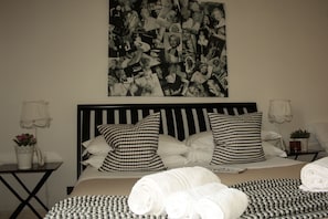 A neatly made bed with checkered pillows, a wall collage of black and white photos, and a bedside table with a lamp and a small plant. - St. John Villa (Rome)