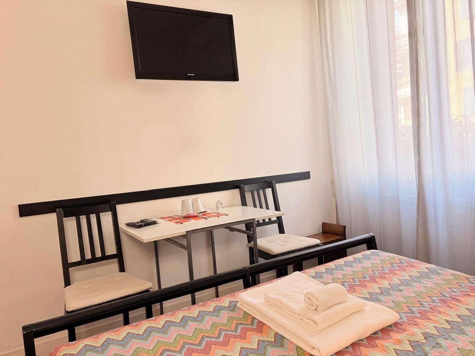 A hotel room with a bed, a desk, a chair, a TV mounted on the wall, and a window with curtains.