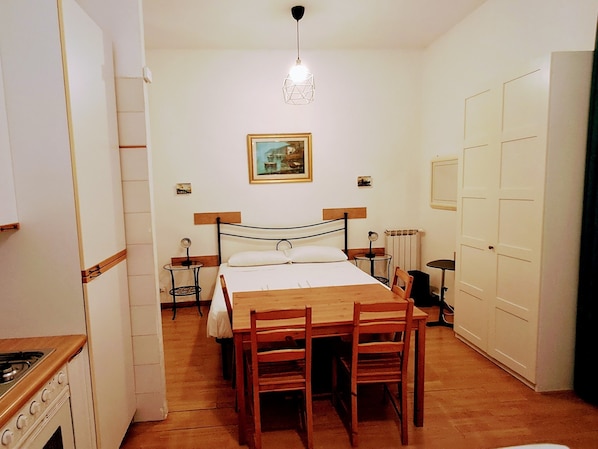 A bedroom with a bed, bedside tables, a wooden table, chairs, a hanging light fixture, and a painting on the wall.