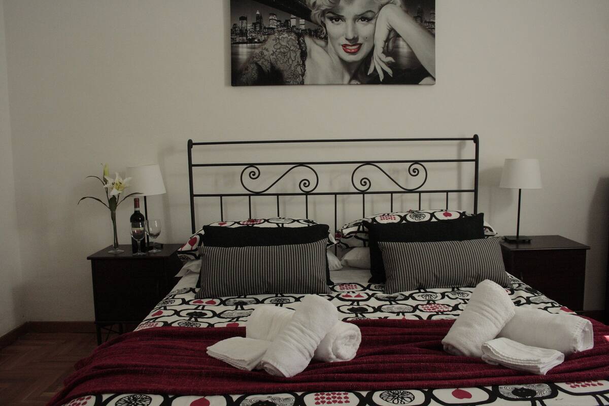 A neatly made bed with a patterned bedspread, two pillows, and a red blanket. There is a nightstand with a lamp and a vase of flowers. A framed picture hangs on the wall above the bed.