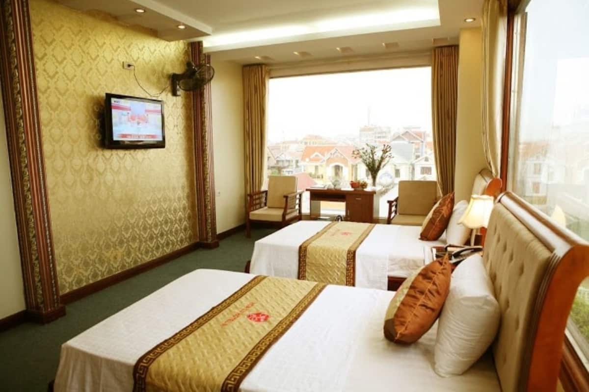 A hotel room with two beds, a television on the wall, a view of buildings outside, and a table with chairs.