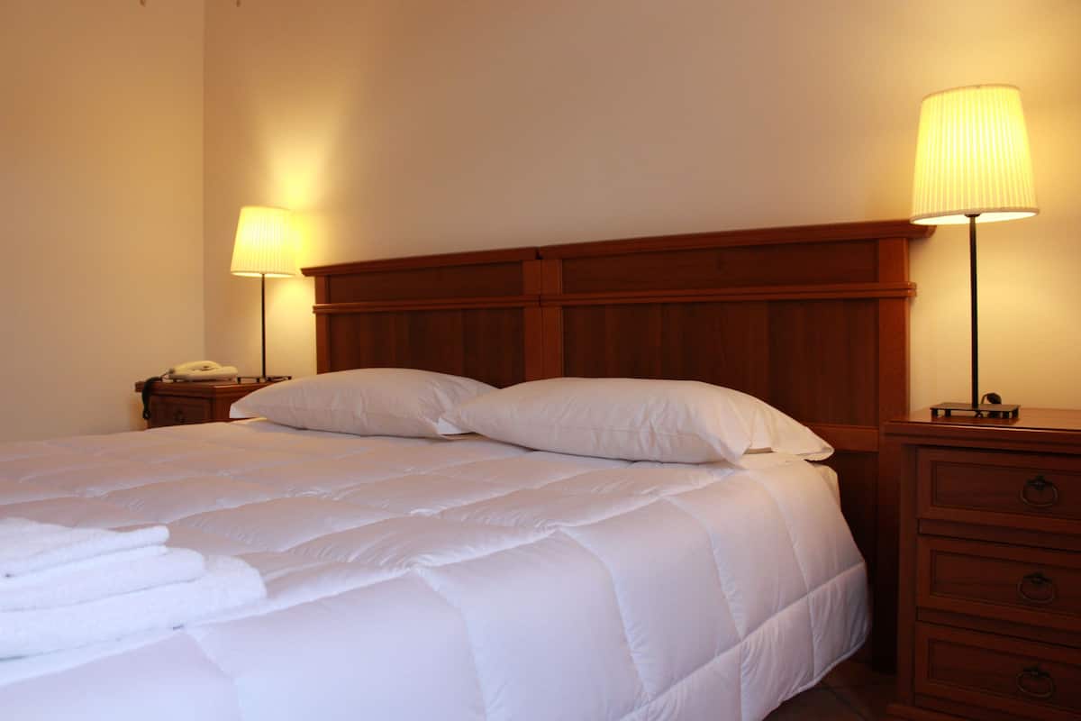 Una cama bien hecha con sábanas y almohadas blancas, flanqueada por dos mesitas de noche con lámparas, en una habitación con cabecera y mesitas de noche de madera.