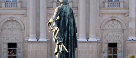 Uma estátua em frente a um edifício imponente com arquitetura clássica, com colunas e janelas em arco.