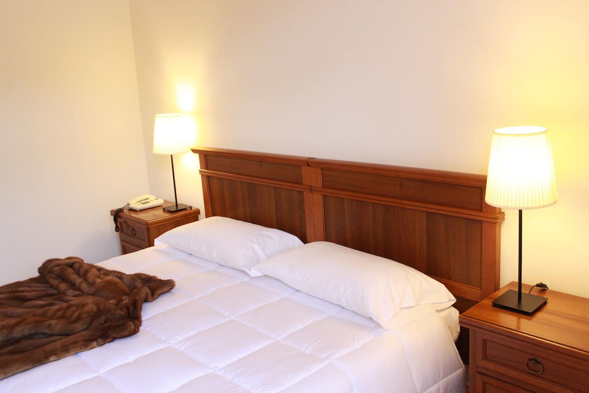 Habitación de hotel con cabecera de madera, cubrecama blanco, dos lámparas de noche, mesita de noche con teléfono y manta marrón.