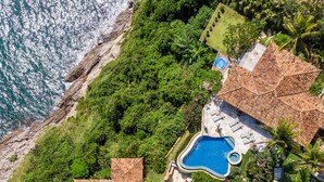 Vista aérea de una propiedad costera con piscina, varias construcciones con techos de tejas y abundante vegetación.